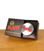 RAW Aluminium Grinder in geöffneter Verpackung, Deckel mit rotem RAW-Logo sichtbar, liegend auf Holztisch.