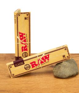 Zwei Packungen RAW Classic Kingsize Slim Rolling Papers, mit Naturfaserband umwickelt, auf einem Stein liegend.