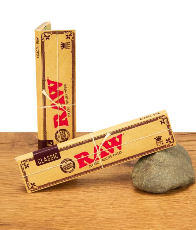 Zwei Packungen RAW Classic Kingsize Slim Rolling Papers, mit Naturfaserband umwickelt, auf einem Stein liegend.