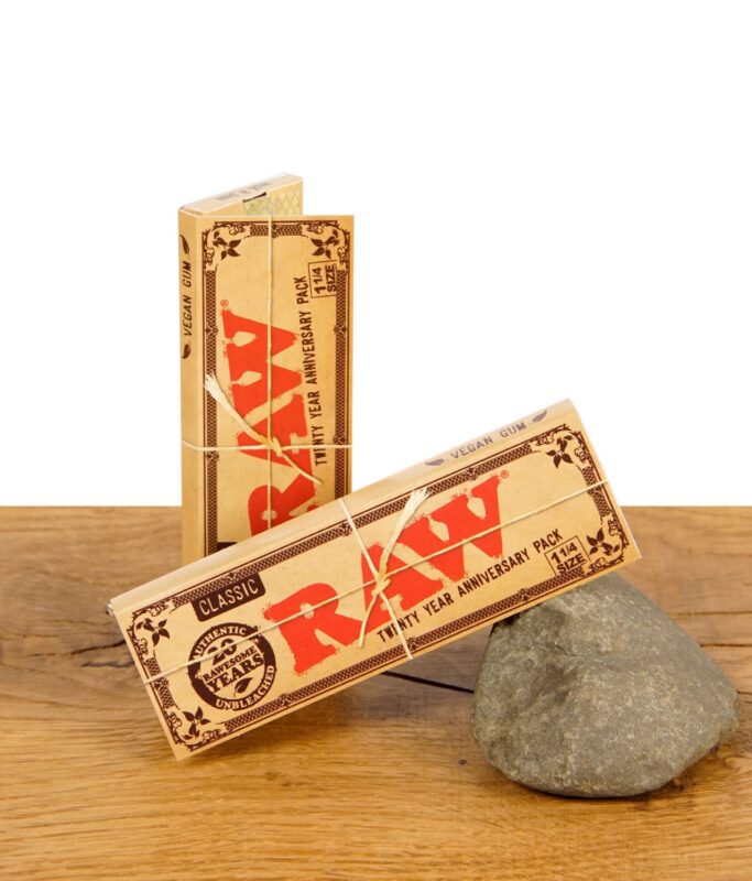 Zwei ungeöffnete RAW Classic Rolling Papers 20th Anniversary Packungen auf Holzuntergrund, eine lehnt an einem Stein