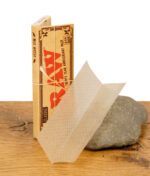 Ein einzelnes ungebleichtes Rolling Paper aus dem RAW Classic 20th Anniversary Pack steht aufgefächert vor der Verpackung