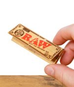 Hand hält eine Packung RAW Classic Rolling Papers 20th Anniversary über einem Holztisch