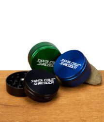 Drei zweiteilige Santa Cruz Shredder Grinder in Schwarz, Blau und Grün auf Holzuntergrund