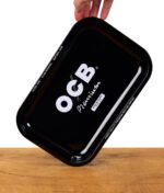 Hand hält ein schwarzes OCB Premium Rolling Tray leicht schräg über einem Holztisch, Fokus auf den weißen Randtext.