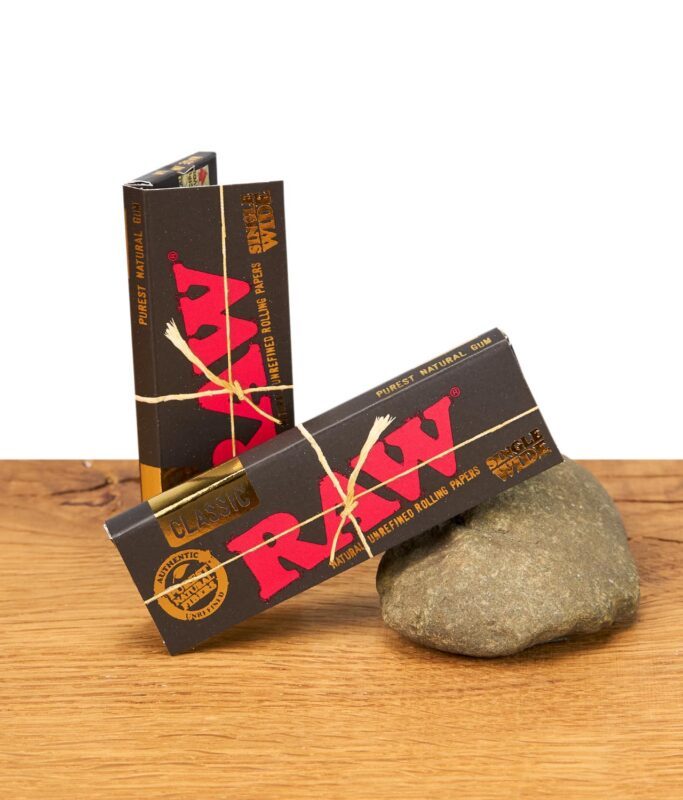 Zwei ungeöffnete Packungen RAW Black Single Wide Papers auf einem Holzuntergrund mit Stein.