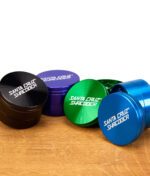 Vier farbige Santa Cruz Shredder Grinder (Schwarz, Lila, Grün, Blau) nebeneinander auf Holzfläche