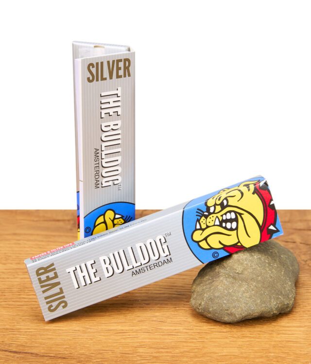 Zwei Packungen The Bulldog Silver King Size Papers + Tips auf Holztisch, eine stehend und eine auf einem Stein liegend