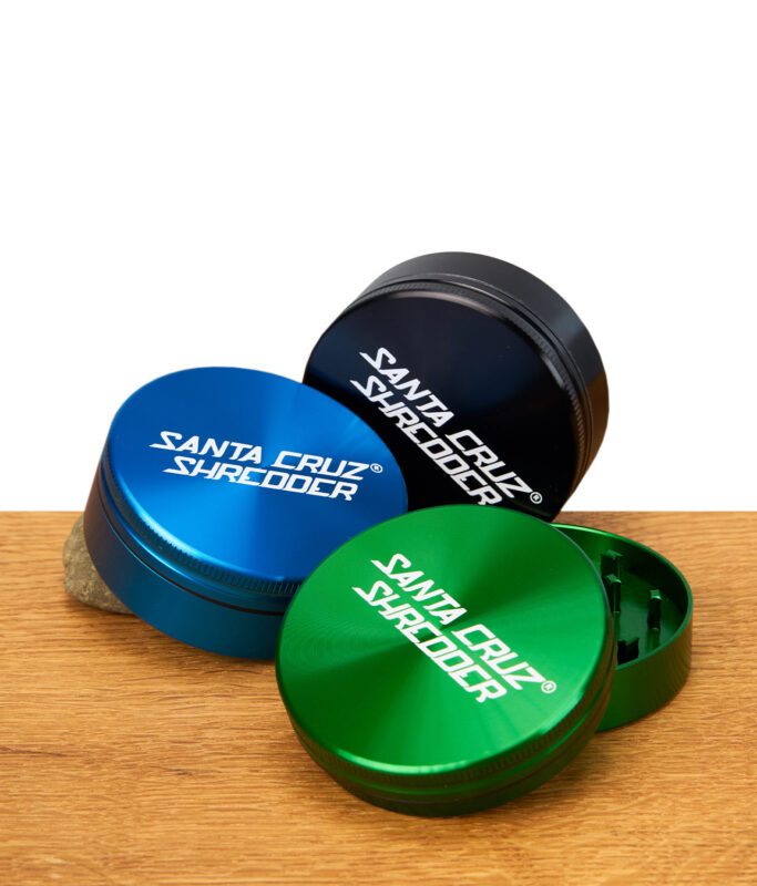 Drei runde Metallmühlen von Santa Cruz in Grün, Blau und Schwarz mit weißem Aufdruck, stehend und liegend auf Holzoberfläche.