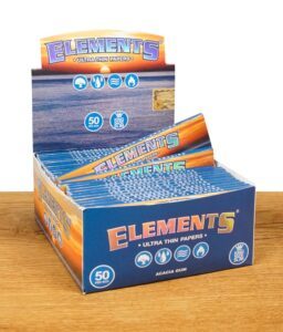 Eine geöffnete Box mit mehreren blauen Verpackungen von Elements Ultra Thin Papers King Size Slim, alle Packungen ordentlich gestapelt auf Holzoberfläche. Das Elements-Logo ist deutlich erkennbar.