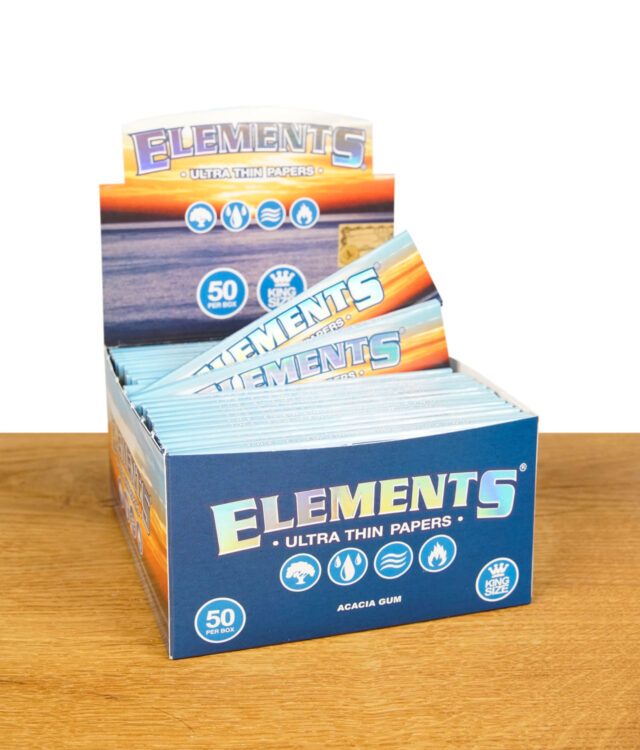 Eine geöffnete Box mit mehreren hellblauen Verpackungen von Elements Ultra Thin Papers King Size, auf Holztisch. Das Elements-Logo ist gut sichtbar.