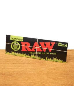 Ein RAW Black Organic Hemp Sticker liegt aufrecht auf einem Holztisch. Zu sehen sind das große rote RAW-Logo, der Text "Organic Hemp Rolling Papers Black" und ein grüner Kreis mit dem Hinweis auf originale Hanffasern.