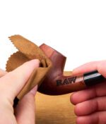 Eine Hand poliert eine RAW Holzpfeife mit einem braunen Reinigungstuch; das RAW-Logo ist auf die Pfeife graviert.