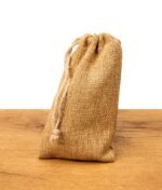 Stehender, geschlossener Jutebeutel mit Zugband auf einem Holztisch, verwendet als Verpackung oder Aufbewahrung.
