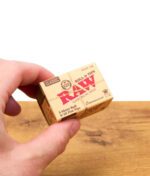 Eine Hand hält eine kleine braune Box RAW Connoisseur Rolls & Tips Classic auf einem Holztisch; auf der Verpackung steht u. a. "5 Meter Roll & 50 Flat Tips".