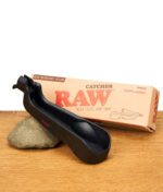 RAW Catcher Aschenbecher aus schwarzem Nylon vor seiner Verpackung, auf einem Stein und einem Holzuntergrund liegend.