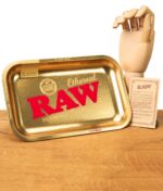 RAW Ethereal Small Rolling Tray in Gold mit rotem RAW-Schriftzug, Classic-Etikett und Produktinfo-Karte, auf einem Holzuntergrund neben einer Deko-Hand.