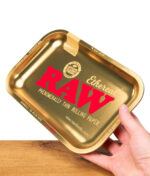 RAW Ethereal Small Rolling Tray, rechteckig und goldfarben, mit großem roten RAW-Logo und Classic-Etikett, wird von einer Hand gehalten, auf Holzoberfläche.