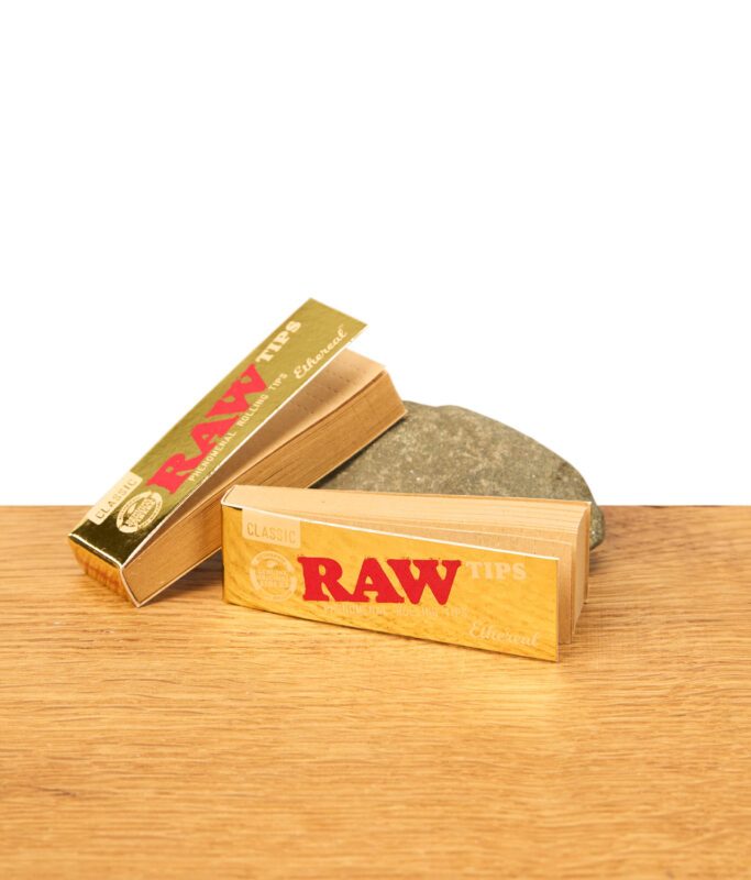 RAW Ethereal Classic Filter Tips, goldene und naturfarbene Verpackungen auf Holz neben Stein, Zubehör für Rolling Papers.