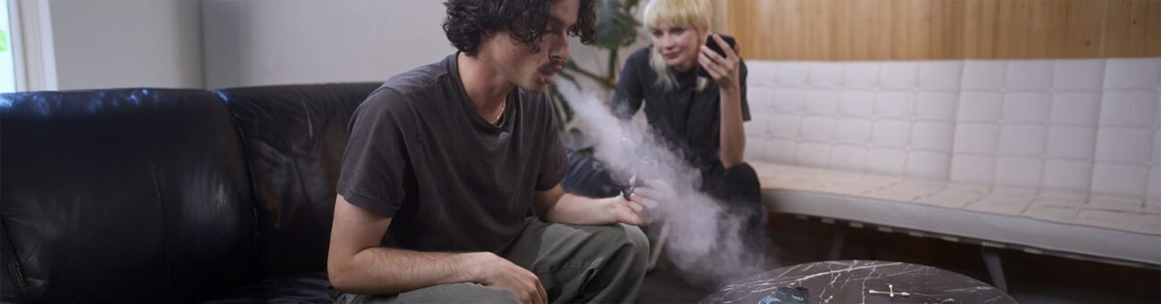 Junger Mann atmet Wolke aus Cannabis Vaporizer mit viel Dampf aus. Frau im Hintergrund schaut zu, in gemütlicher Atmosphäre