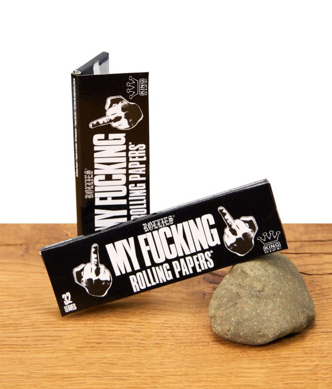 Zwei Packungen Rollies My Fucking Rolling Papers King Size – schwarze Verpackung mit weißem Statement-Design, Drehpapier optimal für Selbstgedrehte, auf Stein platziert.