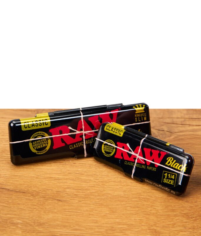 RAW Black Rolling Papers Case Classic King Size Slim und 1 1/4 Size, nebeneinander auf Holztisch – stylische schwarze Metallboxen für Aufbewahrung von Drehpapier.
