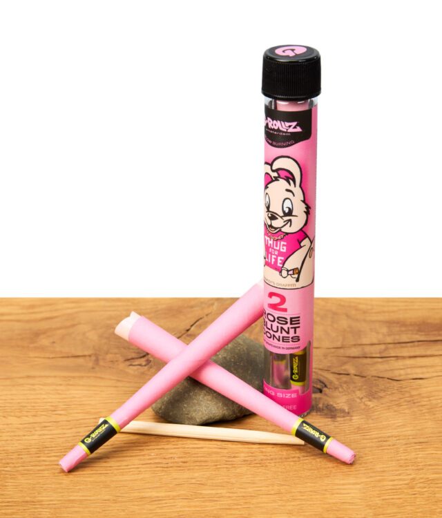 G-Rollz Rose Blunt Cones King Size, zwei pinke Blunt Cones und Verpackung mit Cartoon-Design auf Holztisch – aromatisierte King Size Cones, unverwechselbares Zubehör für Selbstgedrehte.