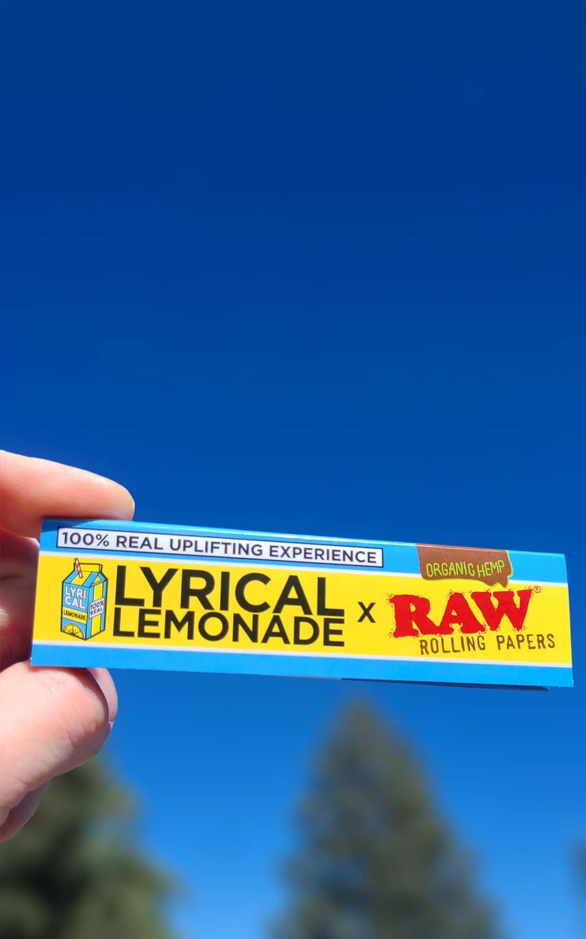 Finger halten Rolling Paper Packung von RAW und Lyrical Lemonade vor strahlend blauem Himmel und Bäumen