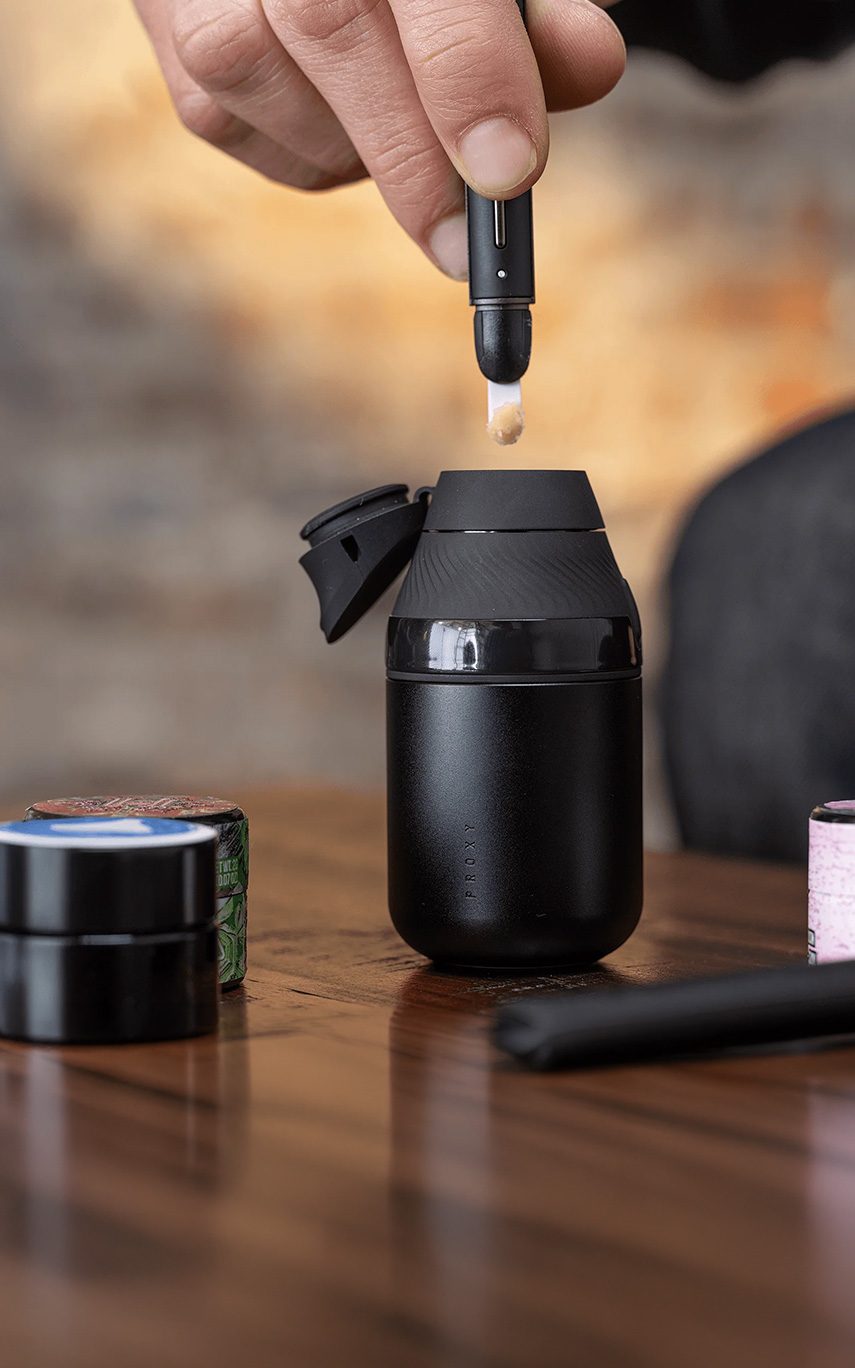 Auf dunklem Holz steht der neue Puffco Proxy Core und wird von oben mit dem Hot Knife V2 von einer Hand beladen.