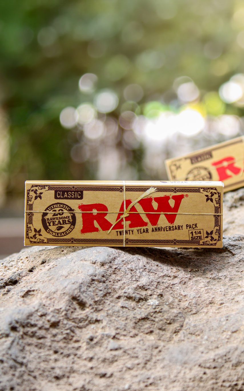RAW Papers kaufen. RAW Rolling Papers Classic Packung auf einem Stein dem klassischen roten Logo. Weitere Packung im Hintergrund zu sehen vor verschwommenem Hintergrund & Bäumen.
