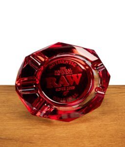 Runder, facettierter RAW Aschenbecher aus rotem Glas steht auf einer Holzoberfläche vor weißem Hintergrund, Draufsicht mit eingeprägtem RAW Schriftzug.