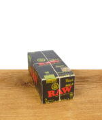 RAW Black Organic Rolls King Size 12er Pack liegt auf einer Holzoberfläche vor weißem Hintergrund, schwarze Verpackung mit rotem RAW-Logo, gelbem „Organic Hemp"-Siegel und weißem Bindegarn.
