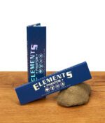 Zwei dunkelblaue Elements Phantom King Size Slim Packungen auf Holzgrund, eine stehend und eine an einen Stein gelehnt.