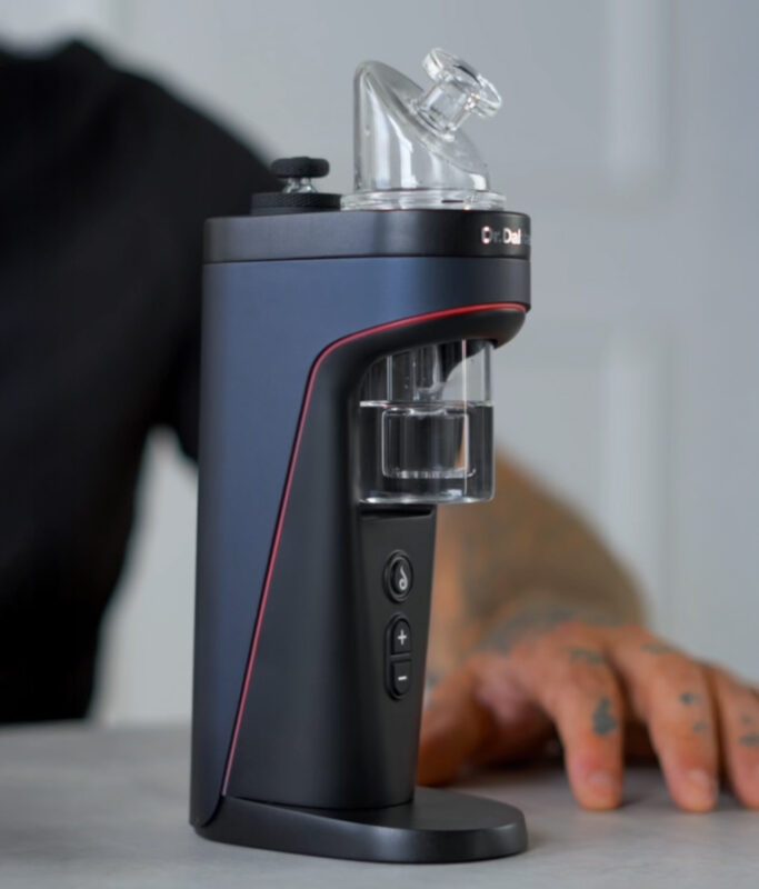 Dr. Dabber Switch 2 Desktop-Vaporizer in Navy Blue mit transparentem Glas-Bubbler-Aufsatz und digitalem Kontrollpanel in einer Lifestyle-Umgebung.