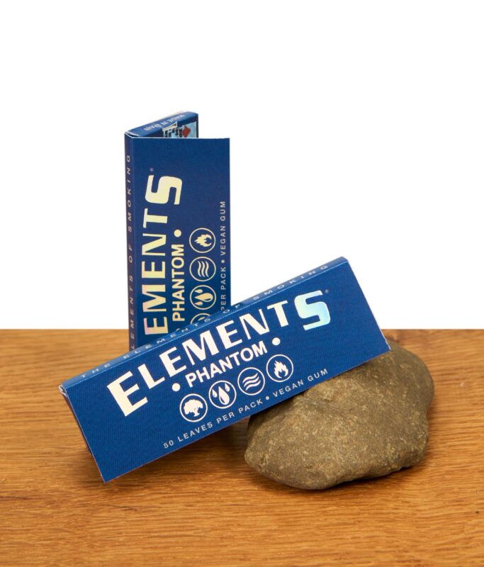Zwei Packungen Elements Phantom Papers 1 1/4 Size mit holografischem Logo auf einer Holzoberfläche neben einem Naturstein.