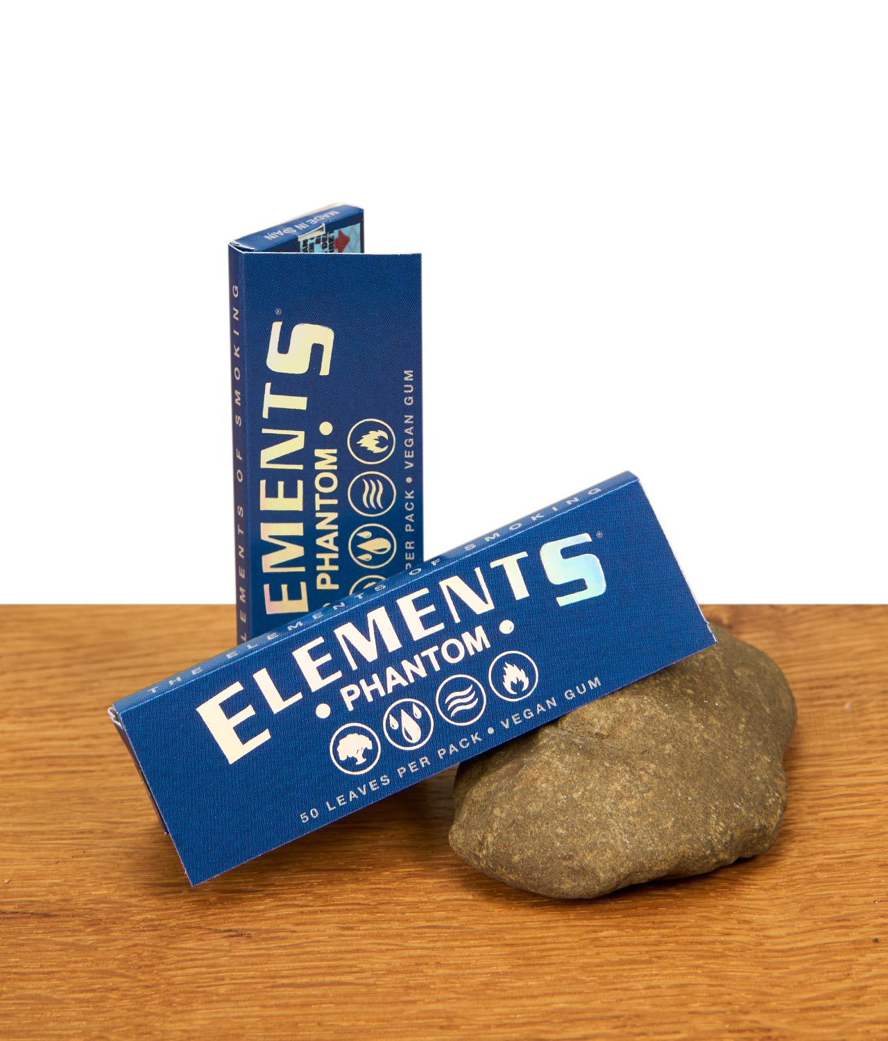 elements-phantom-papers-ein-ein-viertel-size-jpg Zwei Packungen Elements Phantom Papers 1 1/4 Size mit holografischem Logo auf einer Holzoberfläche neben einem Naturstein.