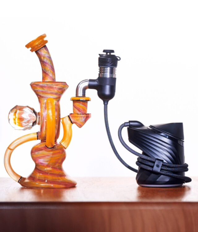Orangene Premium Bong mit Murmel steht auf Holz mit dem schwarzen Puffco Peak Pro. Sie sind mit dem neuen Kabel des Peak Pro Link verbunden. Weißer Hintergrund.