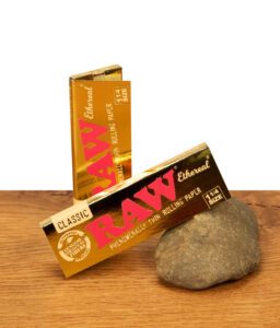 Zwei Packungen RAW Ethereal 1 1/4 Size Papers in glänzender Gold-Optik auf Holzoberfläche mit Naturstein-Akzent.