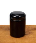 Zylindrische, schwarze Weithalsdose aus Glas mit rundem Schraubdeckel steht auf einer Holzoberfläche vor weißem Hintergrund.