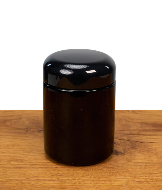 Zylindrische, schwarze Weithalsdose aus Glas mit rundem Schraubdeckel steht auf einer Holzoberfläche vor weißem Hintergrund.