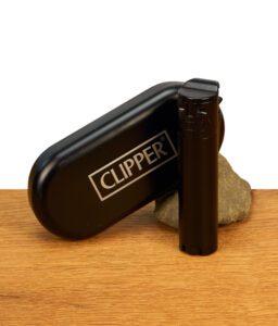 Clipper Feuerzeug Metall Jet-Flame Black mit Reibrad stehend neben passendem Metalletui mit Logo-Prägung auf Holz und Stein.