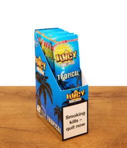 Geöffnetes Display der Juicy Double Wraps Tropical. Die aufgestellte Box auf Holz zeigt mehrere folierte Blunt Wrap Zip-Packs.