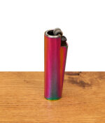 Schrägansicht des nachfüllbaren Metal Clipper Feuerzeugs Rainbow mit sichtbarem Reibradmechanismus, stehend auf einer Holzoberfläche.