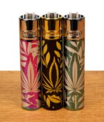 Drei Clipper Metall Jungle Leaves Feuerzeuge mit Hanfblatt-Dekor in Icy-Rainbow, Gold und Silber auf einer Holzoberfläche.