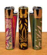 Drei Clipper Metall Jungle Leaves Feuerzeuge in Icy-Rainbow, Gold-Schwarz und Silber-Grün mit Hanfblatt-Dekor auf Holzoberfläche.