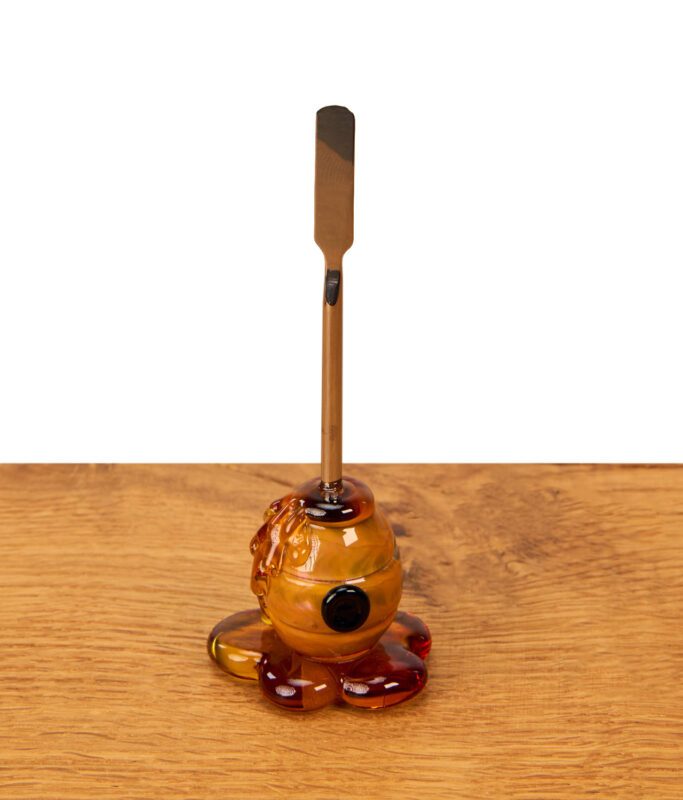 Empire Glassworks Honey Dabber auf einem Holztisch. Das Dabbing Tool hat einen flachen Metallspatel und einen Honigtopf-Griff aus Glas.