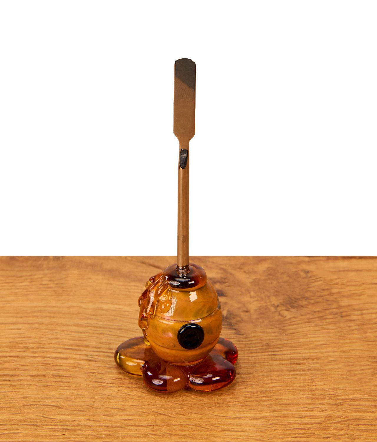 dabbin-tool-honey-dabber-von-empire-glassworks-jpg Empire Glassworks Honey Dabber auf einem Holztisch. Das Dabbing Tool hat einen flachen Metallspatel und einen Honigtopf-Griff aus Glas.