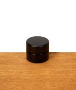 Geschlossene Miron Weithalsdose 5ml aus blickdichtem Violettglas mit schwarzem Schraubdeckel auf einer Eichenholz-Oberfläche.