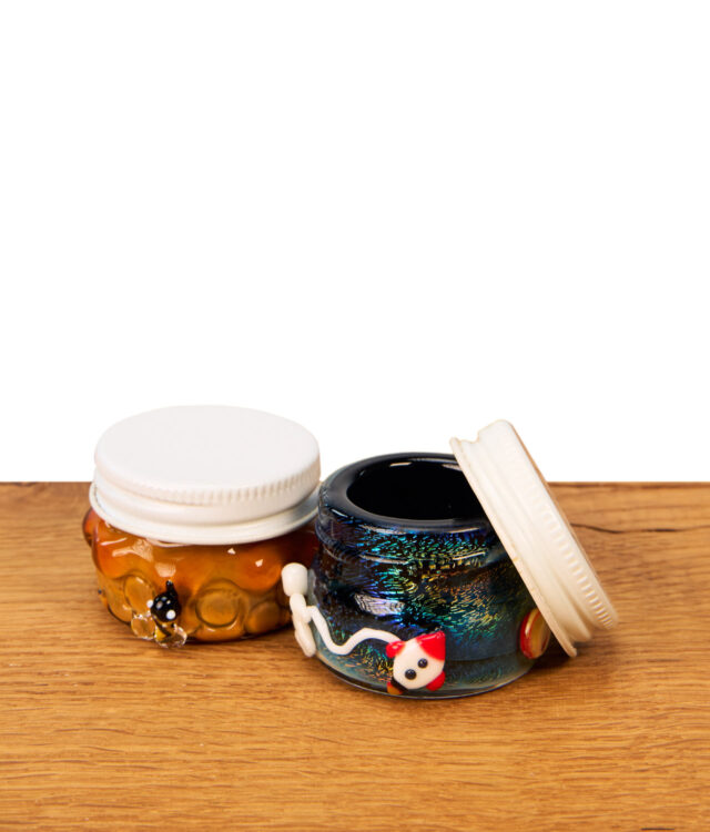 Zwei Empire Glassworks Terp Jars auf Holztisch. Aufbewahrungsgläser mit 3D-Glasapplikationen, rechts geöffnet mit weißem Schraubdeckel.