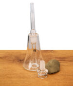 Der MJ Arsenal Lumina Bubbler aus klarem Borosilikatglas mit seiner charakteristischen konischen Form; davor liegt das passende gläserne Köpfchen auf einer Holzoberfläche.