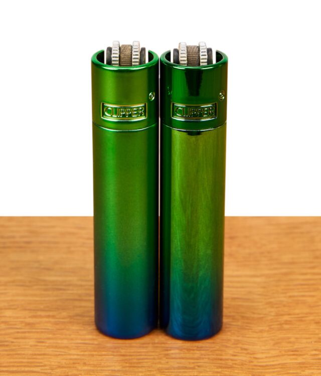 Clipper Metall Peacock Feuerzeuge im 2er-Set mit mattem und glänzendem Grün-Blau-Farbverlauf nebeneinander auf Holz.
